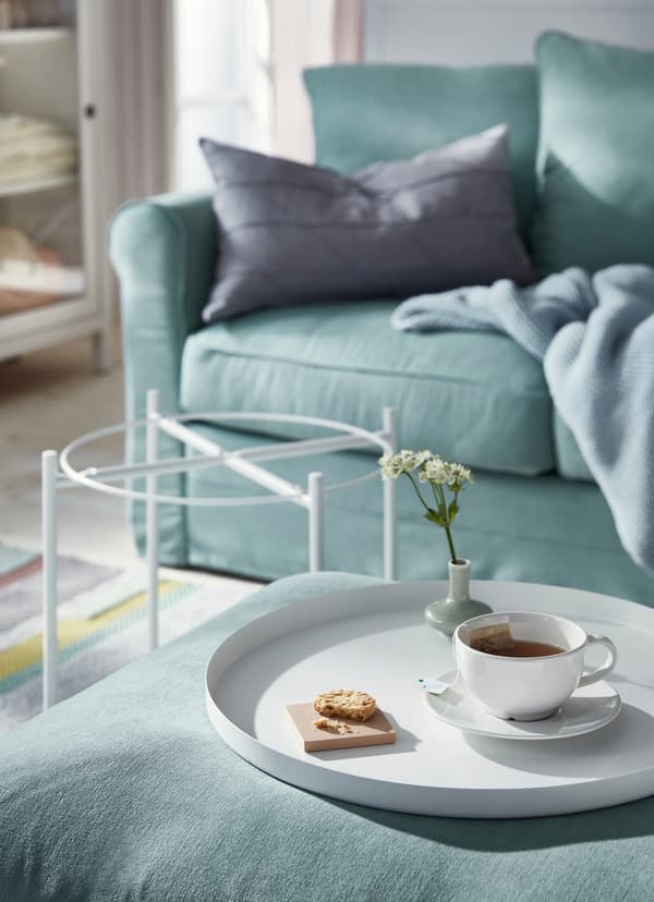 Tasse de thé et biscuit sur le plateau blanc de la table plateau blanche IKEA GLADOM, posé sur le repose-pieds d’un canapé vert.