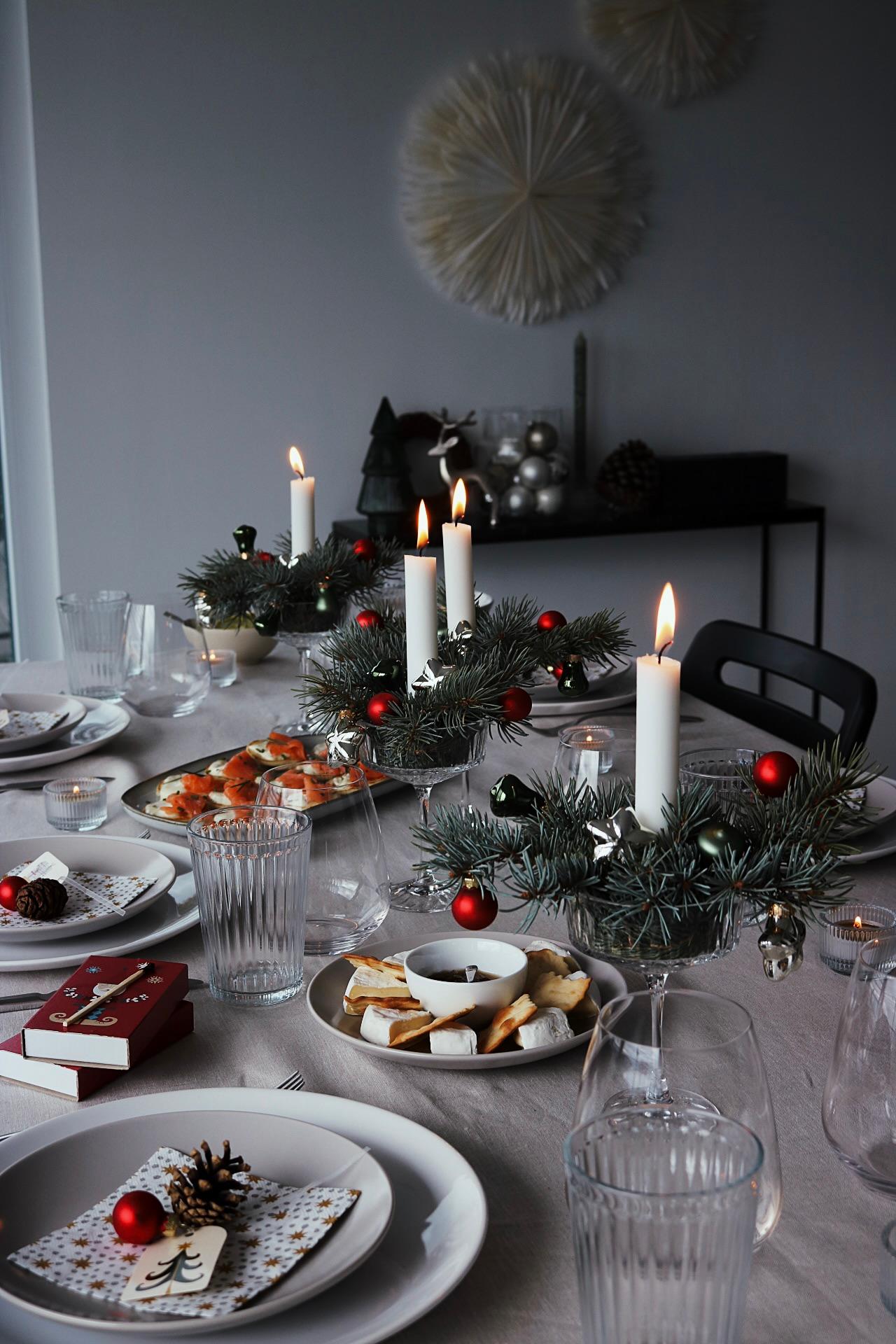 Tavola imbandita a festa con candele bianche accese, centrotavola sempreverdi, decorazioni rosse e un'elegante mise en place. Atmosfera festiva accogliente e invitante.