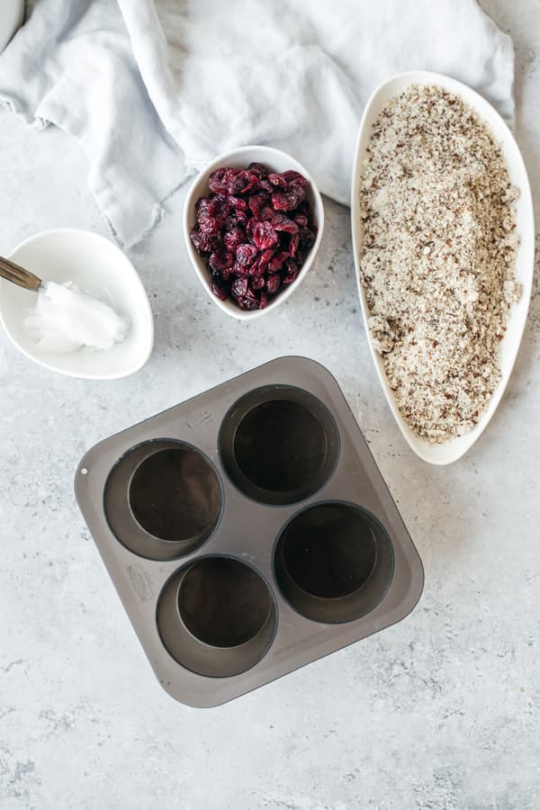 Teglia da muffin, ciotole con mirtilli rossi, farina e riso su superficie chiara, accessori da forno sullo sfondo.