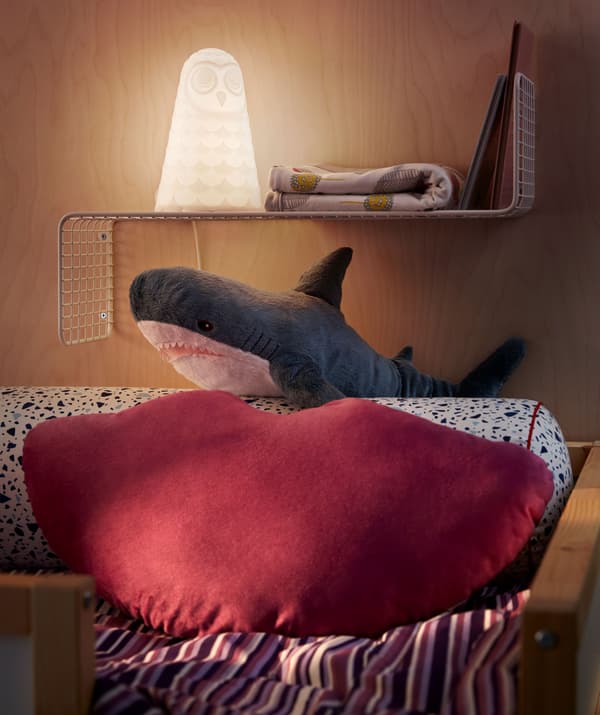 Tête du lit haut garnie d’un coussin et d’une peluche requin, une étagère murale étroite fait office de table de chevet et accueille veilleuse, livres et couverture.