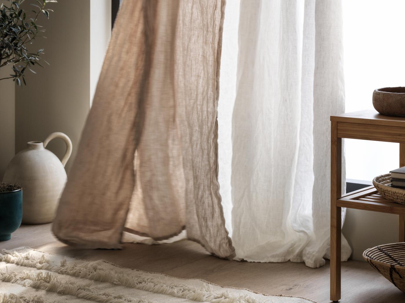 The bottom of a beige curtain and a sheer white SILVERLÖNN curtain touch a wooden floor and billow in front of a window.