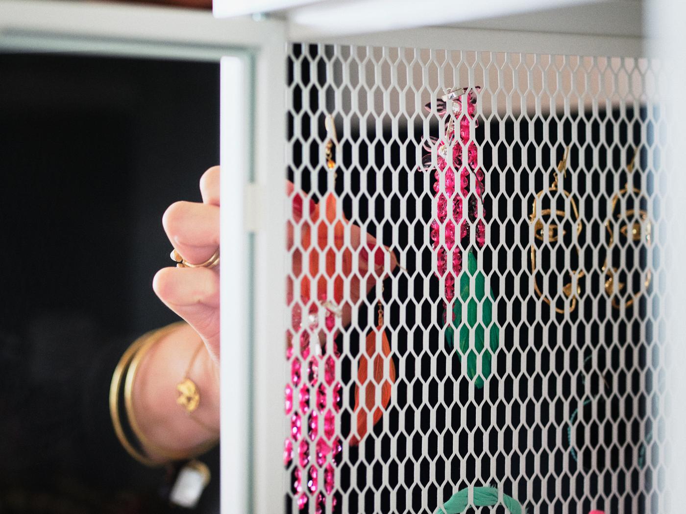 The hand of a person wearing gold bracelets and rings opening the white mesh metal door of a BAGGEBO cabinet.