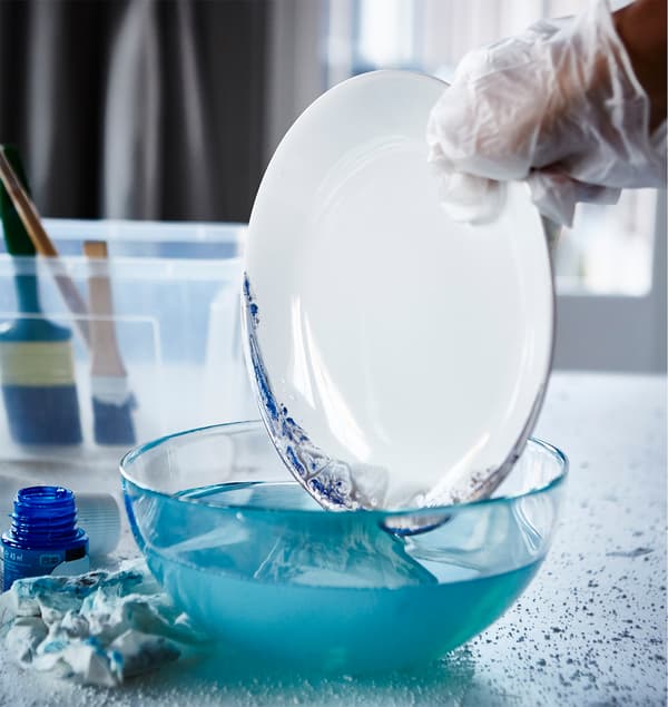 The newly-painted edge of an IKEA FÄRGRIK plate is being dipped into a bowl of water.
