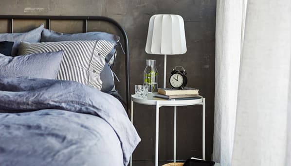 The side of a made bed with a bedside table, glass of water, books, an alarm clock and dimmable lamp