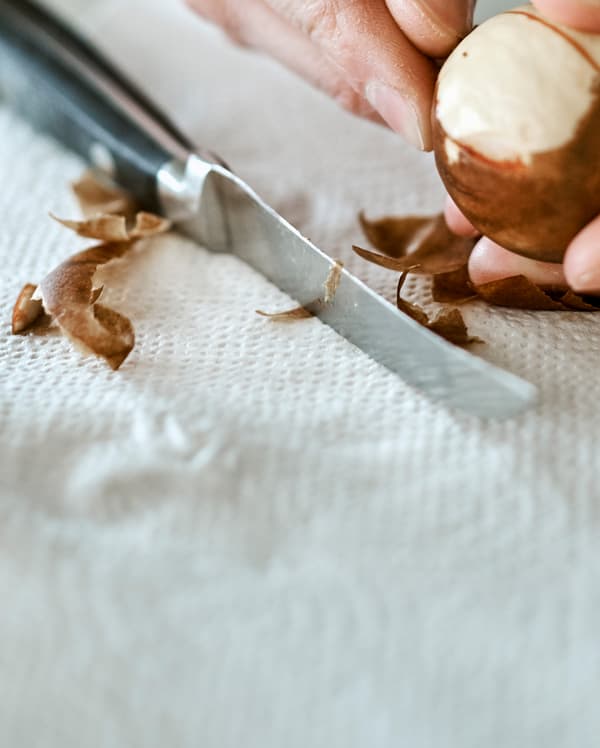 The skin being peeled from an avocado seed with the peel on a paper towel next to a kitchen knife with a black handle.