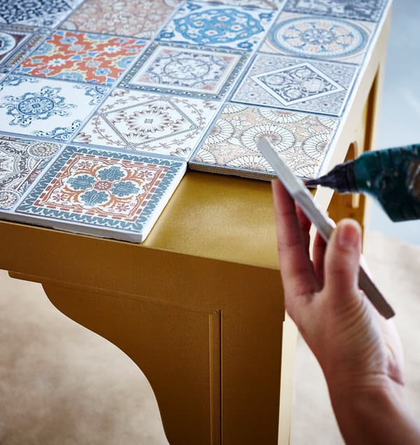 The top of a gold IKEA LACK table is being decorated with a variety of patterned tiles.