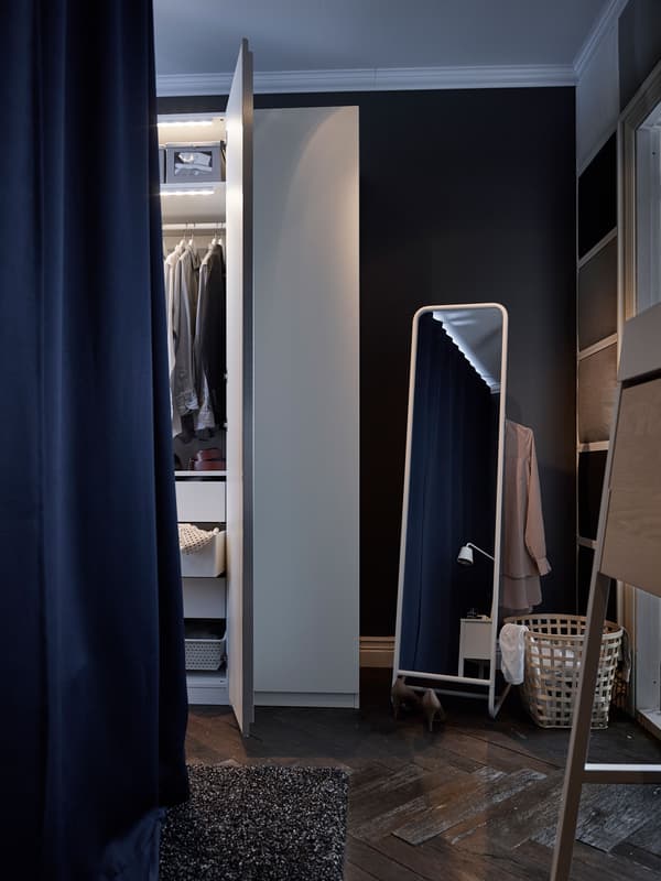 This white bedroom wardrobe with built-in lighting stands next to a mirror with built-in clothes storage.