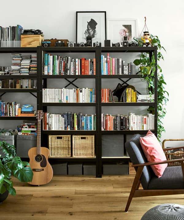 Three black bookcases filled with colour co-ordinated books, wicker baskets, framed photos, vintage cameras and plants.