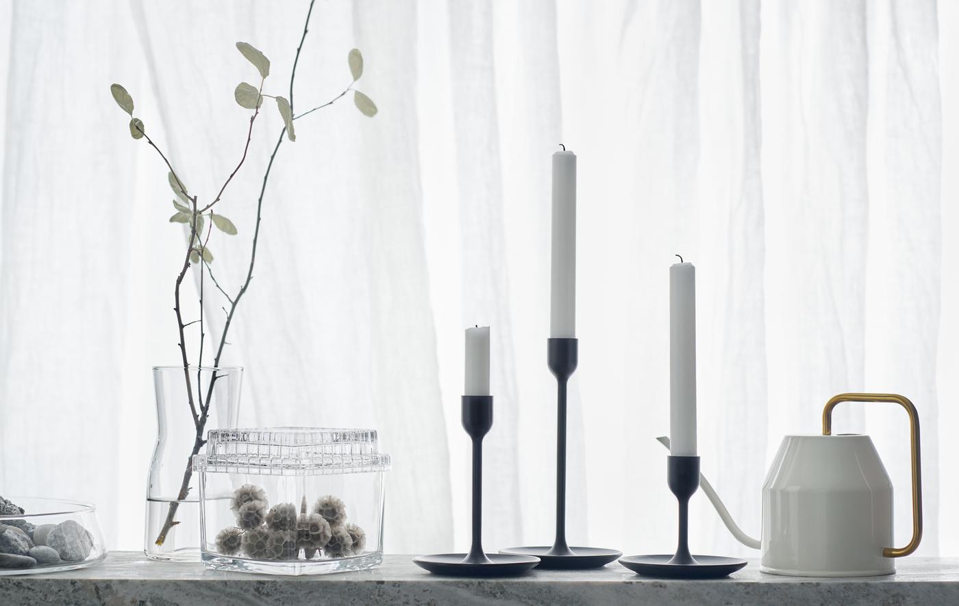 Three black candlesticks of different heights on a white backdrop with watering can and glass jars.