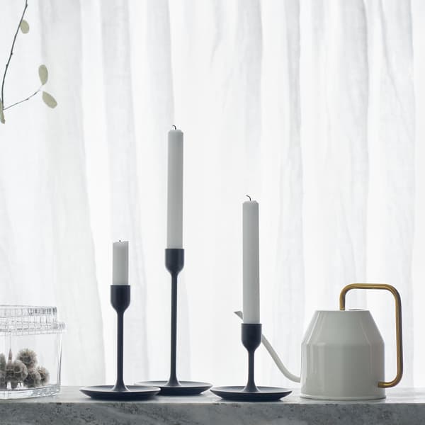 Three black candlesticks of different heights on a white backdrop with watering can and glass jars.