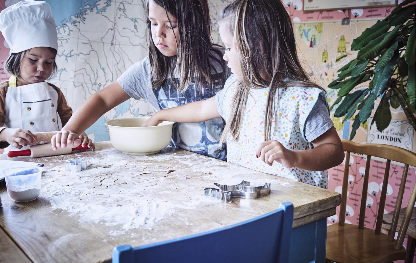 Three children standing around a table with flour, dough and baking utensils.