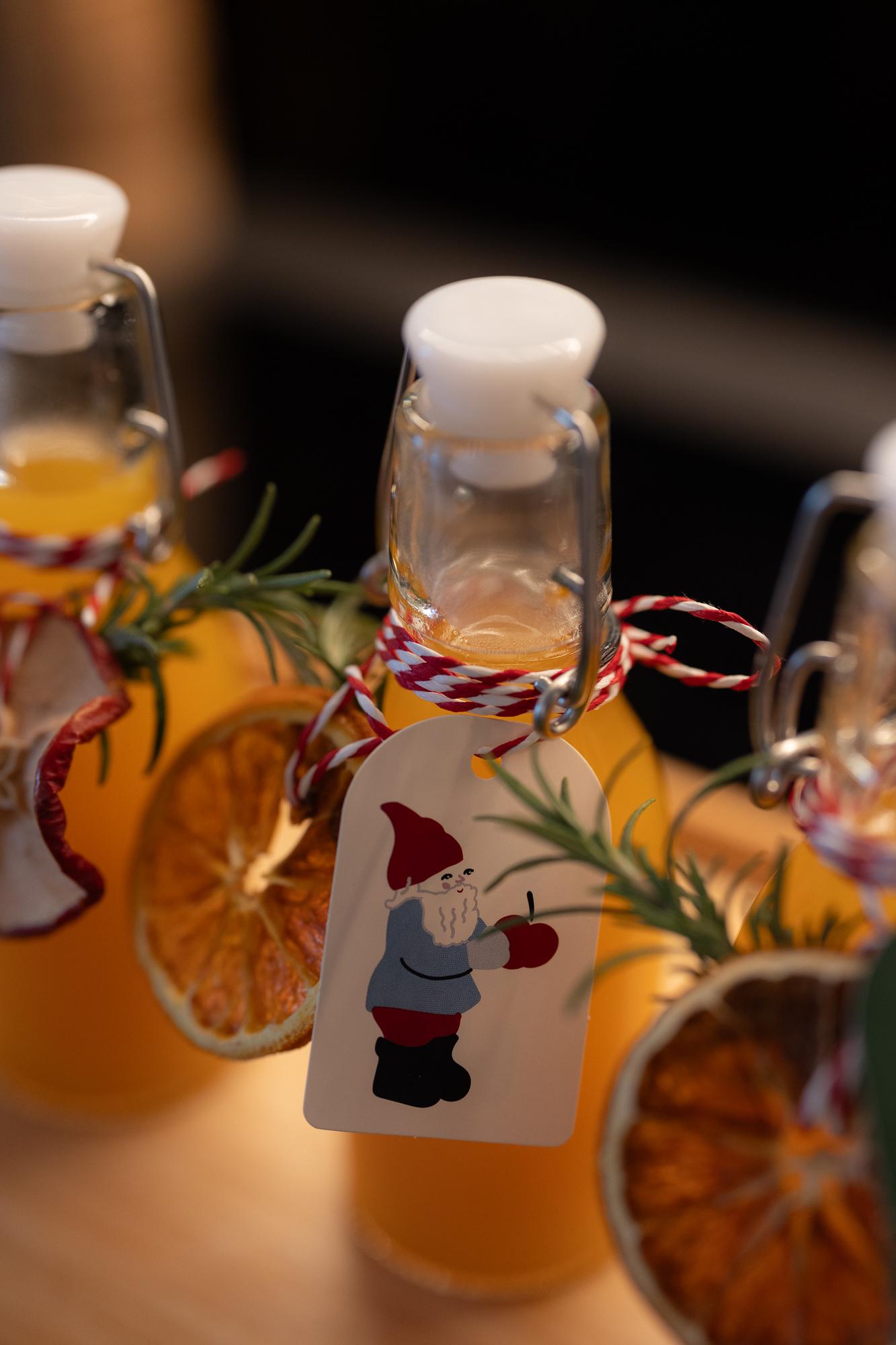 Three glass bottles filled with a golden drink, decorated with orange slices, rosemary, and festive tags featuring a gnome.