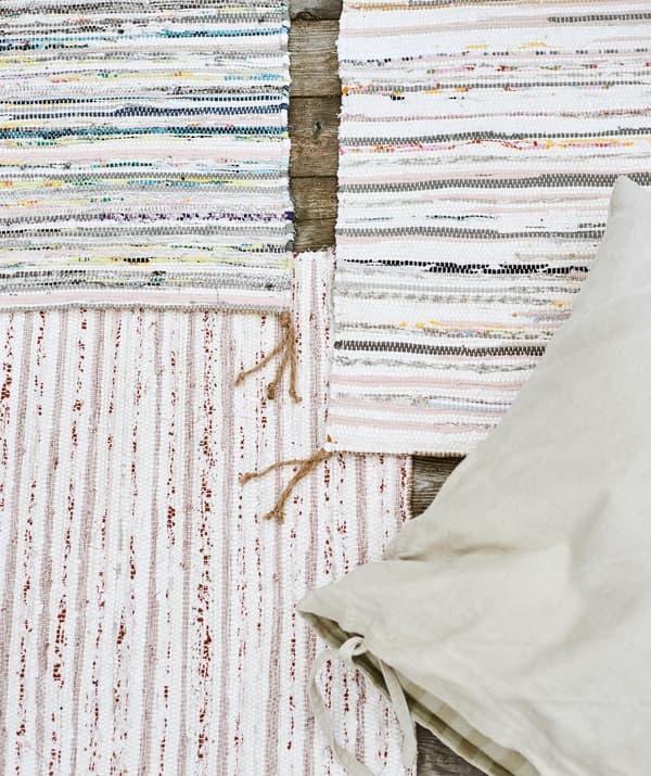 Three handwoven multicoloured striped rugs overlapping each other on wood decking, with a beige linen cushion on top.