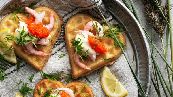 Three heart shaped VÅFFLOR waffles with shrimps, mayonnaise, seaweed pearls and dill on a plate with a kitchen towel.