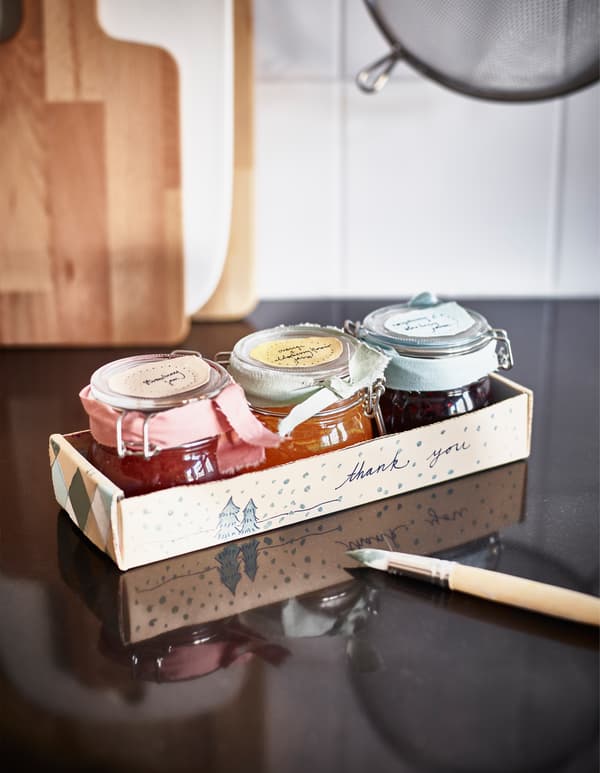 Three jars of jam decorated with ribbon in a cardboard tray that’s been painted with a thank you.