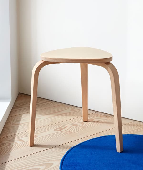 Three-legged, light-wood stool standing in a sharp-angle corner on a wooden floor, one leg on a small blue rug.
