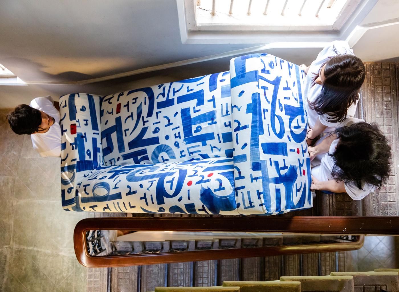 Three people with dark hair carrying a blue and white KLIPPAN sofa up the stairs.