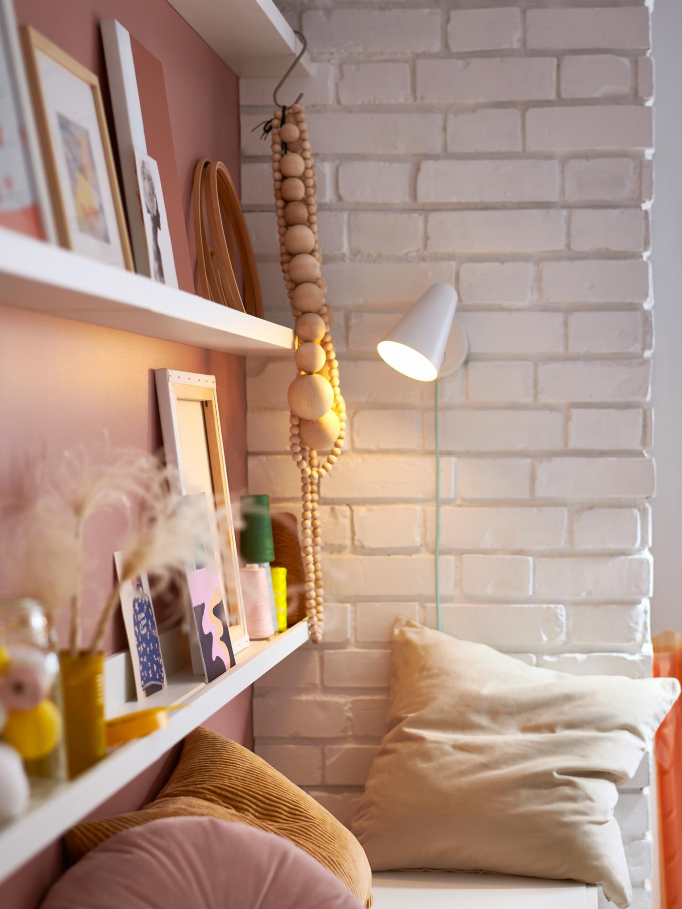Three rows of white MOSSLANDA picture ledges, framed pictures, accessories, a wall lamp and a white bench with cushions.