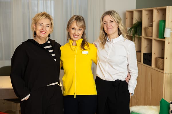 Three women smile at the camera while standing in a newly furnished room. An IKEA employee in a yellow jacket stands in the middle.