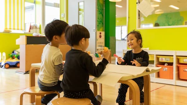 Three young children sitting at a low table in a bright playroom, drawing on large paper with toys and storage units in the background