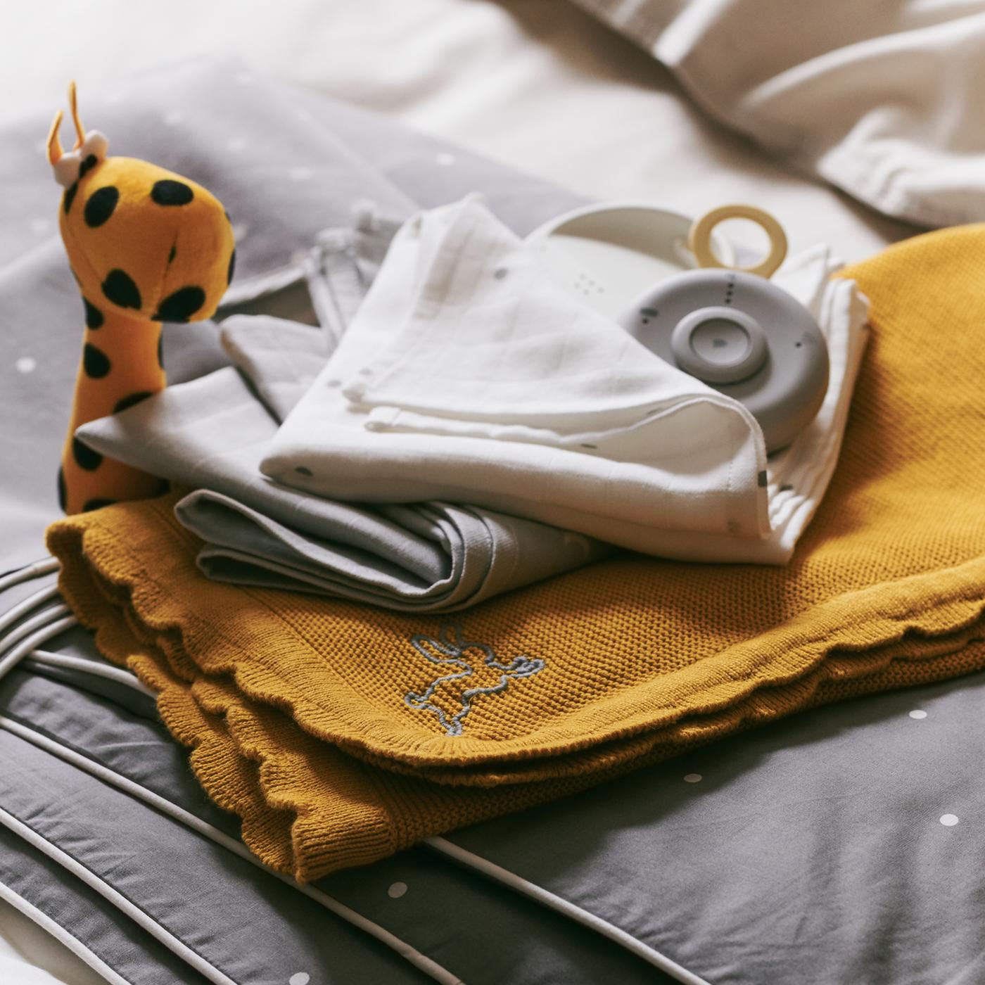 Tindanan tekstil bayi termasuk selimut SOLGUL berwarna kuning gelap dan kuilt katil bayi LENAST, dengan pengawas bayi UNDVIKA terletak di atasnya.