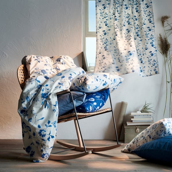 Tissu à motif floral bleu et blanc déposé sur un fauteuil berçant en rotin, un rideau présentant le même motif habille la fenêtre en arrière-plan.