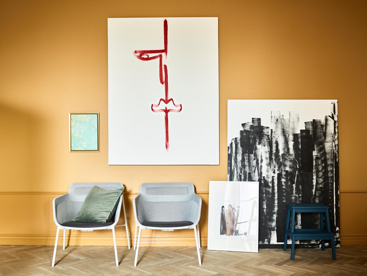 Toiles avec des coups de pinceau rouges et noirs accrochées sur un mur ocre ou posées contre celui-ci. Deux chaises se trouvent devant.