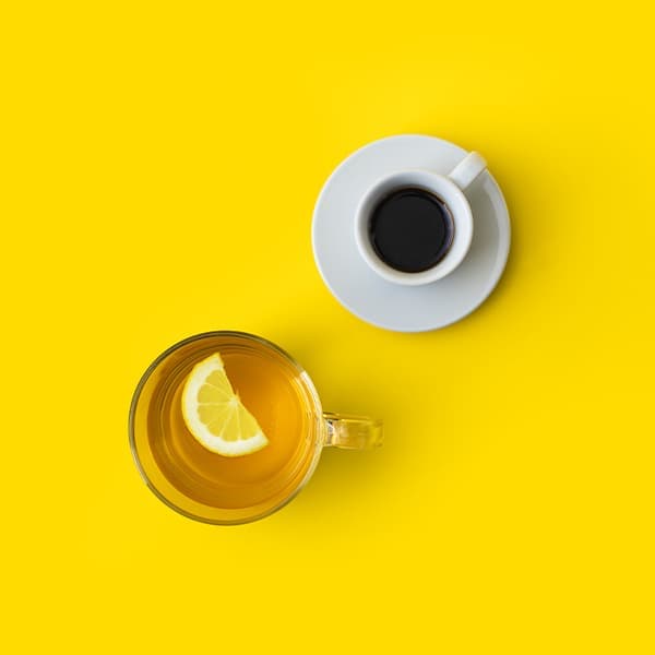 Top view of a white cup of black coffee on a saucer and a clear glass cup of tea with a lemon slice, both placed on a bright yellow background.