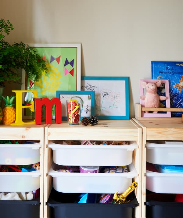 Toys stored in plastic trays in a wooden frame, with plants, pictures and books on top.