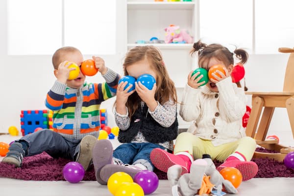 Tres niños, dos niñas y un niño jugando en un piso blanco con juguetes de colores. Se tapan los ojos con pelotas de colores de materiales responsables para que jueguen con seguridad en casa. Hay pelotas azules, moradas, amarillas, verdes y naranjas, con un par de juguetes de madera de fondo también.