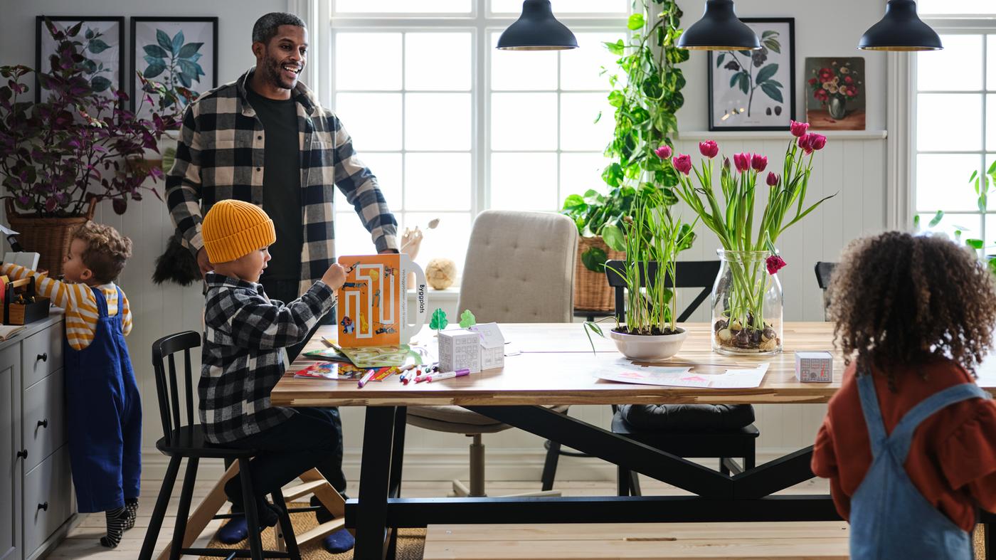 Trois jeunes enfants et leur père s’occupent joyeusement dans la zone salle à manger avec une table SKOGSTA.