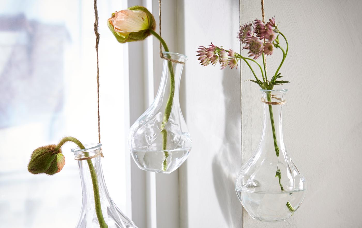 Trois vases en verre garnis d’une fleur chacun, accrochés à une fenêtre pour une nature morte colorée.