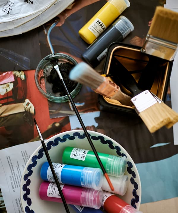 Tubes de peinture dans un bol blanc et pinceaux dans une boîte noir et or sur une table recouverte de papier journal.