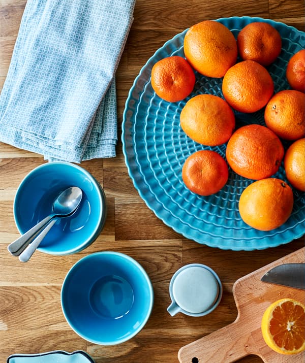Turquoise teacups, napkins and a serving plate with satsumas on a wooden worktop next to a chopping board and cut satsuma.