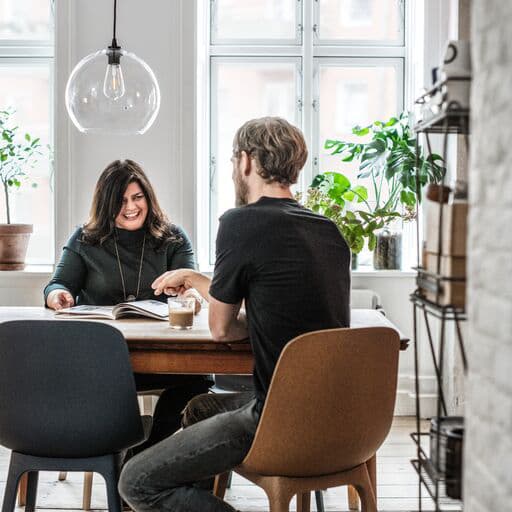 Två personer sitter vid ett träbord i ett ljust rum med stora fönster och krukväxter; en läser, den andra dricker kaffe.

