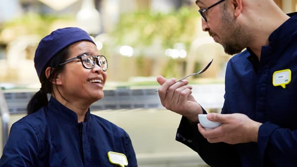 Twee IKEA medewerkers in donkerblauwe uniformen eten een snack en lachen.