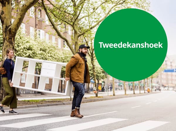 Twee personen dragen samen een wit opbergmeubel over een zebra­pad in een stadsstraat met bomen, naast een groen rond label met de tekst “Tweedekanshoek”.
