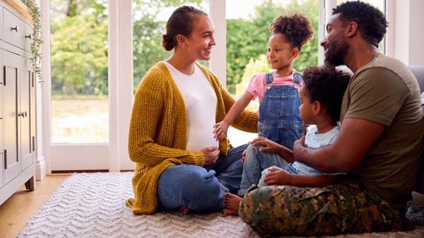 Two adults and two children sit on a rug indoors, smiling and talking together near a bright window.