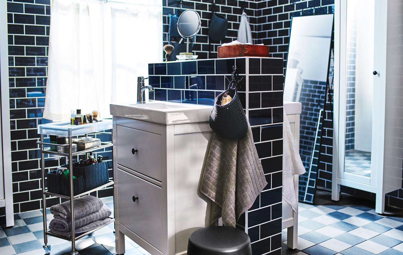 Two bathroom sinks and washstands mounted back to back in the centre of a large, black tiled bathroom.
