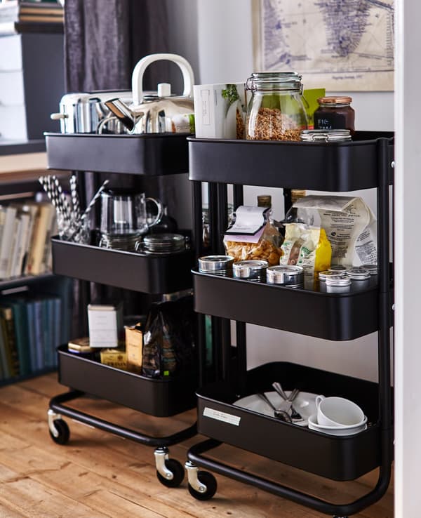 Two black RÅSKOG kitchen trolleys stocked with kitchen supplies, including food, a toaster, a kettle, and dishes.