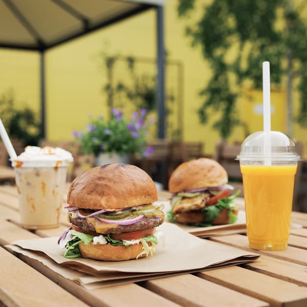 Two burgers on a wooden table outdoors, served with iced coffee and peach-mango smoothie.
