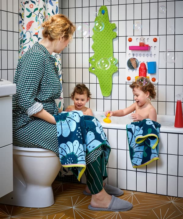 Two children in a white tiled bath and a woman sitting on the lid of a toilet next to them.