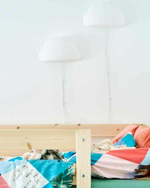 Two cloud-shaped children’s wall lights fixed to the wall beside a wooden bed frame, the bed has colorful bedding.