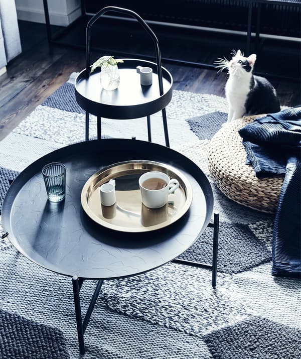 Two dark side tables and a rattan footstool on a grey textured rug.