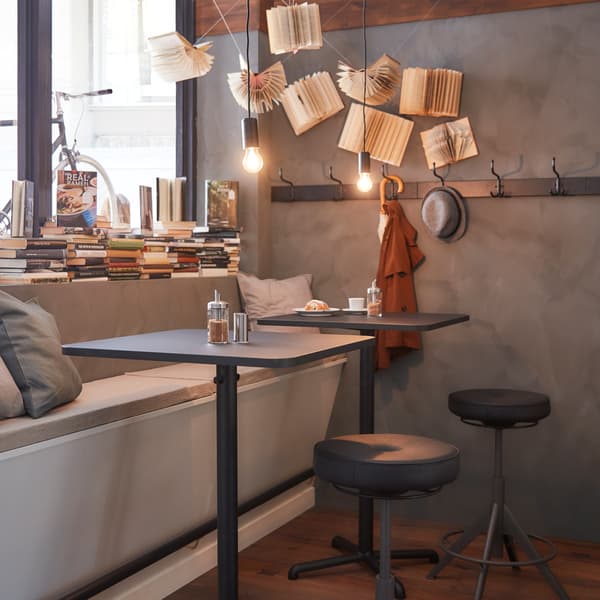 Two dark square tables next to a window with books in it, a bench with grey pillows and two black sit and stand supports.