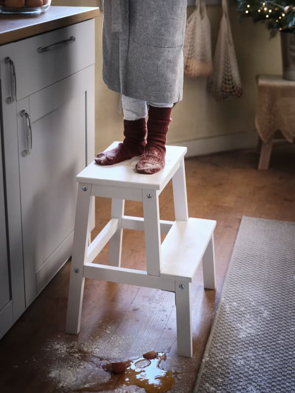 Two feet covered in white flour are stood on a BEKVÄM step stool in a traditional-looking kitchen.