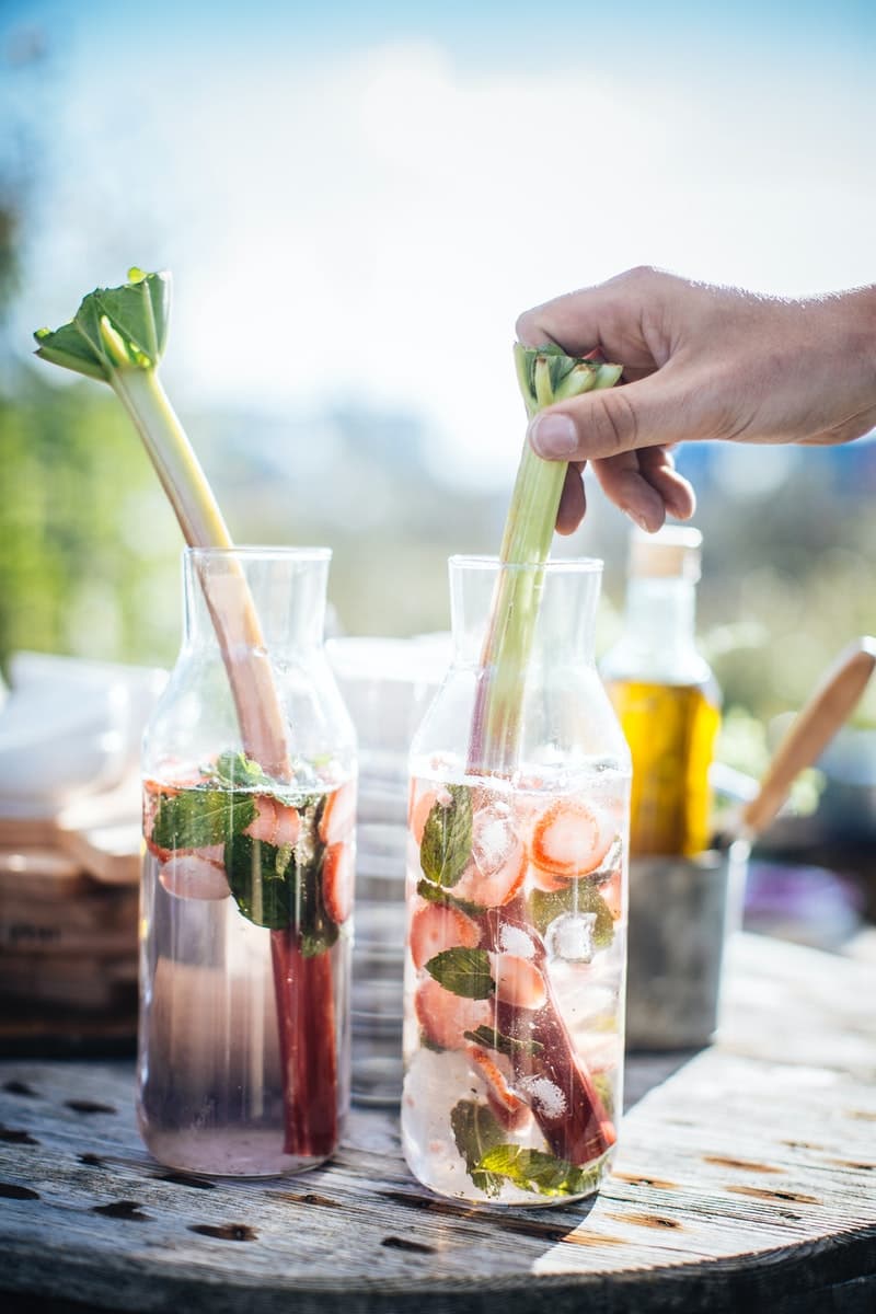 Two glass bottles filled with a drink containing strawberries, mint leaves, and rhubarb stalks are on a table, while a hand holds one of the rhubarb stalks in the right-hand bottle.