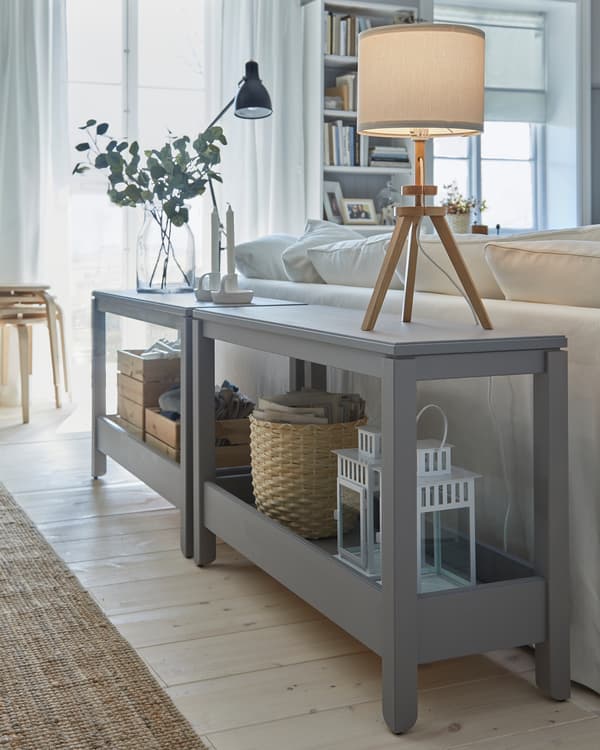 Two grey console tables are placed behind a sofa and serve as decorative room dividers. Two table lamps are placed on top.