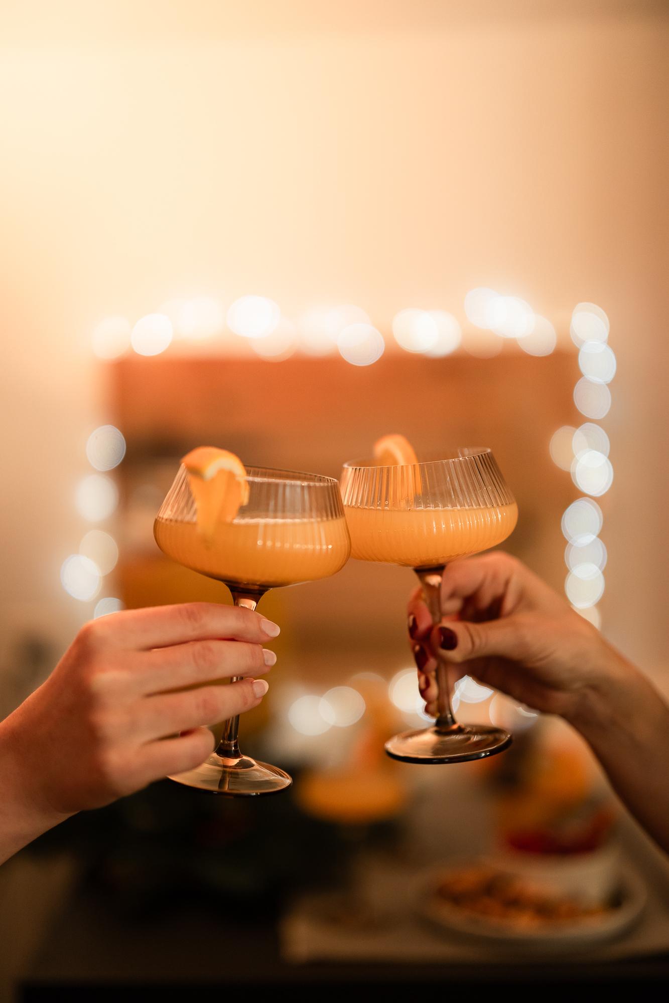 Two hands raise elegant glasses of orange cocktails, clinking them together against a softly lit background.