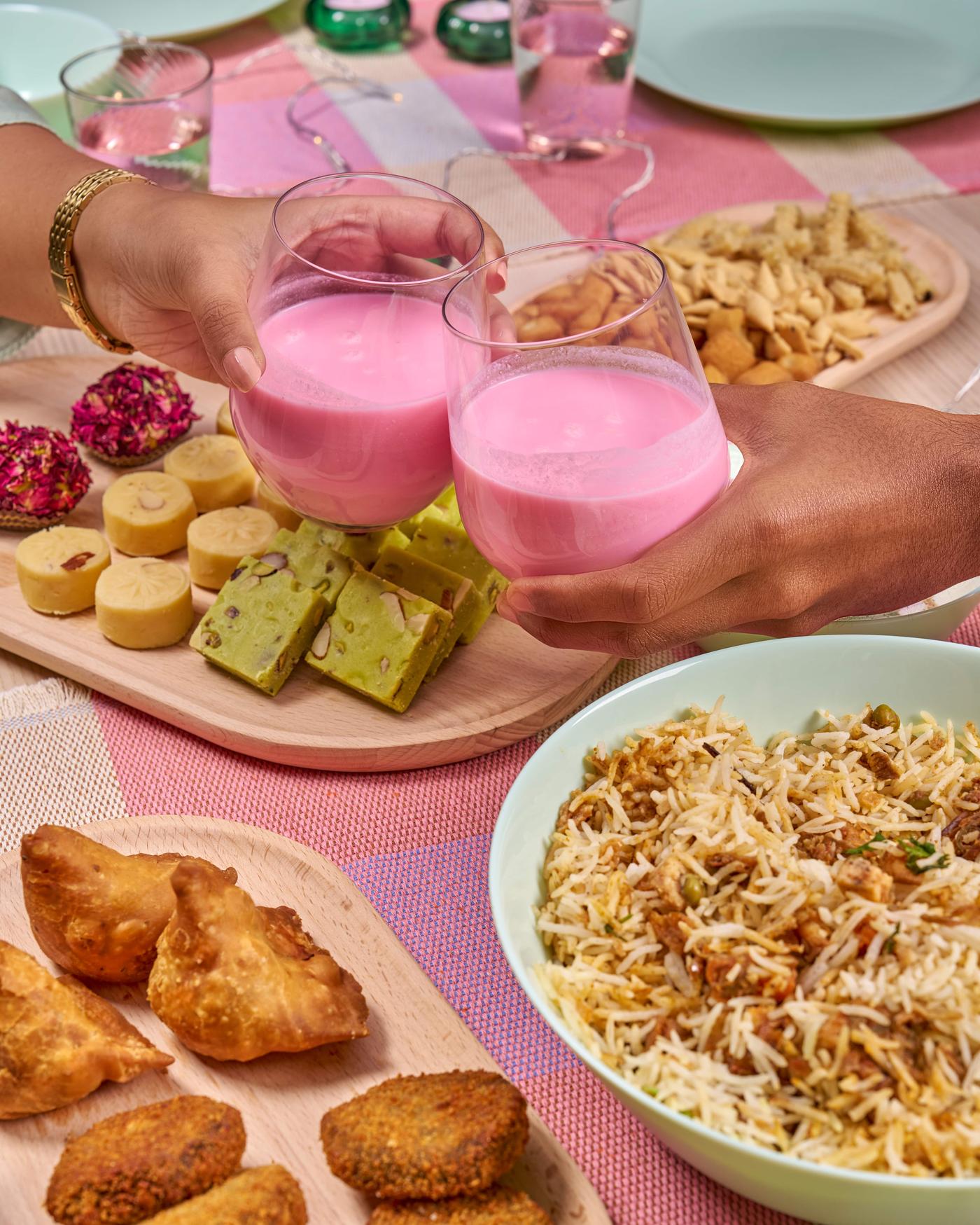 Two hands raising glasses of pink rose milk sharbat for a toast, with sweets and snacks on the table.
